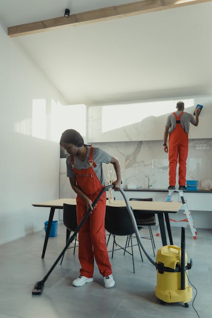 Two professional cleaners from West Hampstead Cleaner are engaged in surface cleaning within a modern, well-lit residential kitchen or dining area. The cleaner in the foreground is using a vacuum or s