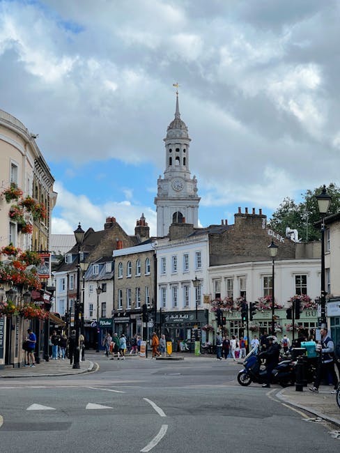 A lively street scene in West Hampstead featuring a mix of historic and modern buildings, with a prominent white clock tower rising above the rooftops under a partly cloudy sky. The street is bustling with pedestrians walking along the pavements, some window shopping and others engaging in conversation. Several small shops and cafes line the street, their windows displaying various goods and outdoor seating areas. In the foreground, a motorbike is parked near a black lamppost adorned with hanging flower baskets. The scene is well-lit with natural daylight, highlighting the architectural details and vibrant activity, characteristic of West End Lane in West Hampstead. This image captures the charm and community atmosphere of the area, suitable for content related to local urban life and community spaces.