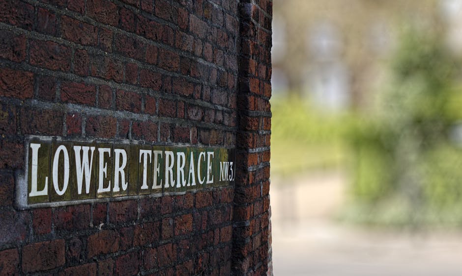 A close-up view of a brick wall featuring a rectangular metal street sign with white uppercase letters reading 'LOWER TERRACE NW3' mounted on the dark red brick surface. The bricks show a weathered texture with some mortar visible between them. The background is blurred with hints of greenery, indicating an outdoor setting. The image emphasizes the brick texture and clear signage, typical of residential or urban street scenes, and is provided by West Hampstead Cleaner as part of their surface cleaning and deep cleaning services for end of tenancy in West Hampstead NW6.