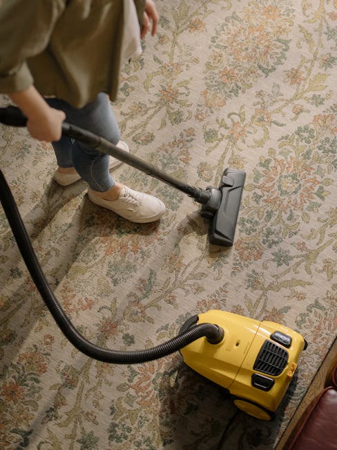 A person using a vacuum cleaner with a yellow canister and black hose to clean a patterned area rug carpeted floor in a room. The flooring features a floral and geometric design in muted colors, with natural light illuminating the carpet. The individual is dressed casually in light-colored trousers and white sneakers, and is operating the vacuum head over the surface. The scene highlights domestic surface cleaning and stain removal, with visible attention to maintaining cleanliness, consistent with services offered by West Hampstead Cleaner for deep cleaning and sanitisation of carpets in residential spaces.